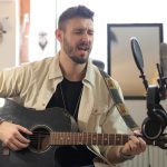 Músico canta e toca violão acústico em um estúdio de gravação.