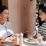 Duas mulheres elegantemente vestidas conversam durante uma refeição em um café ao ar livre.