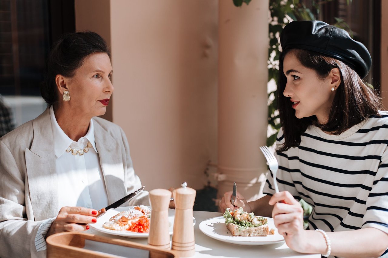 Duas mulheres elegantemente vestidas conversam durante uma refeição em um café ao ar livre.
