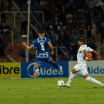 Dois jogadores disputam intensamente a bola durante uma partida de futebol, com torcedores ao fundo.