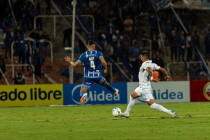 Dois jogadores disputam intensamente a bola durante uma partida de futebol, com torcedores ao fundo.