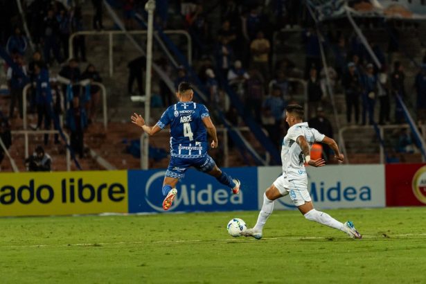 Dois jogadores disputam intensamente a bola durante uma partida de futebol, com torcedores ao fundo.