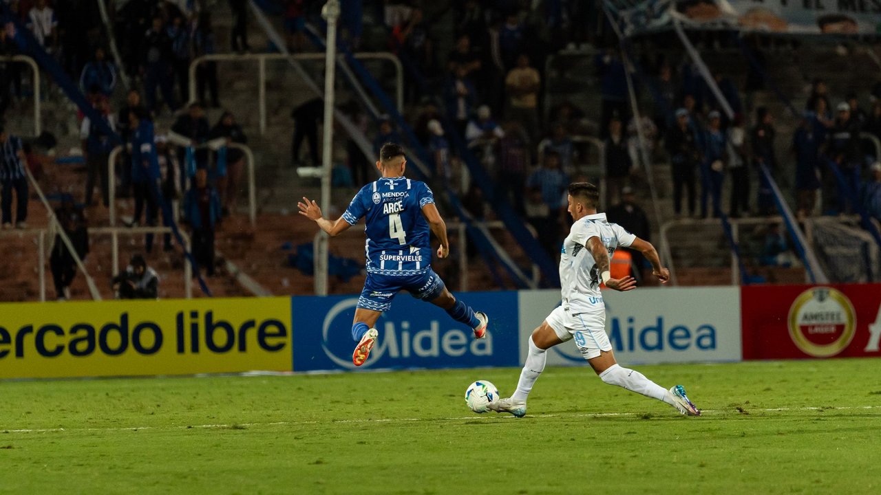 Dois jogadores disputam intensamente a bola durante uma partida de futebol, com torcedores ao fundo.