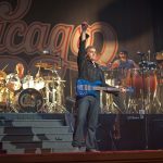 Músicos se apresentam em um palco durante um show, com bateria, guitarra e baixo.