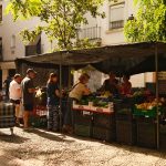 Feira de rua movimentada, com pessoas comprando produtos frescos em um ambiente urbano ensolarado.