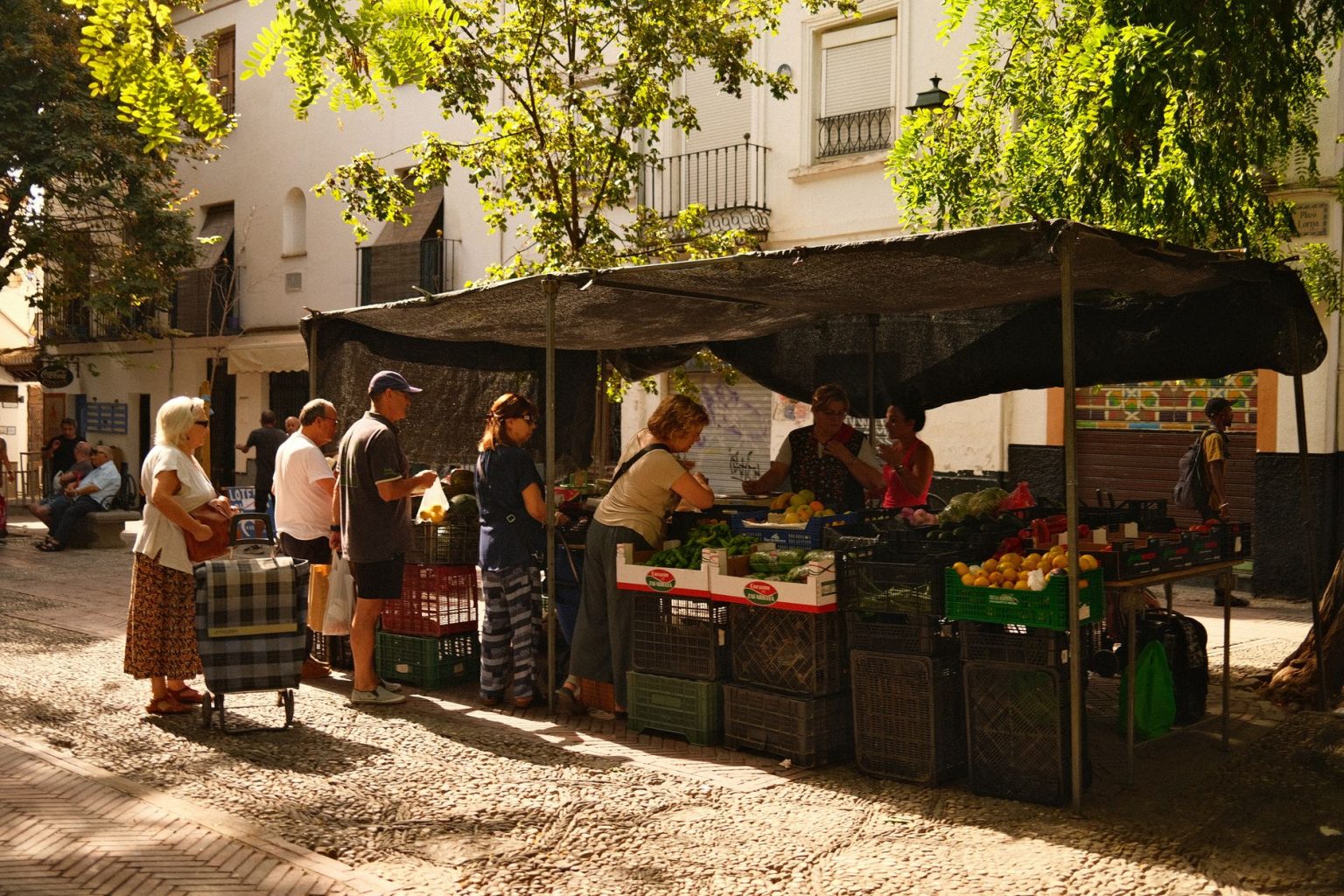 Feira de rua movimentada, com pessoas comprando produtos frescos em um ambiente urbano ensolarado.