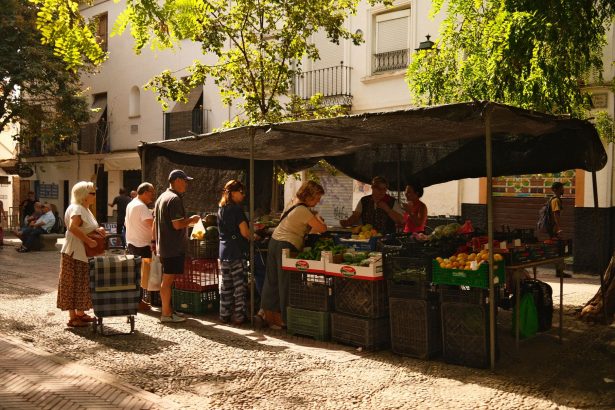 Feira de rua movimentada, com pessoas comprando produtos frescos em um ambiente urbano ensolarado.
